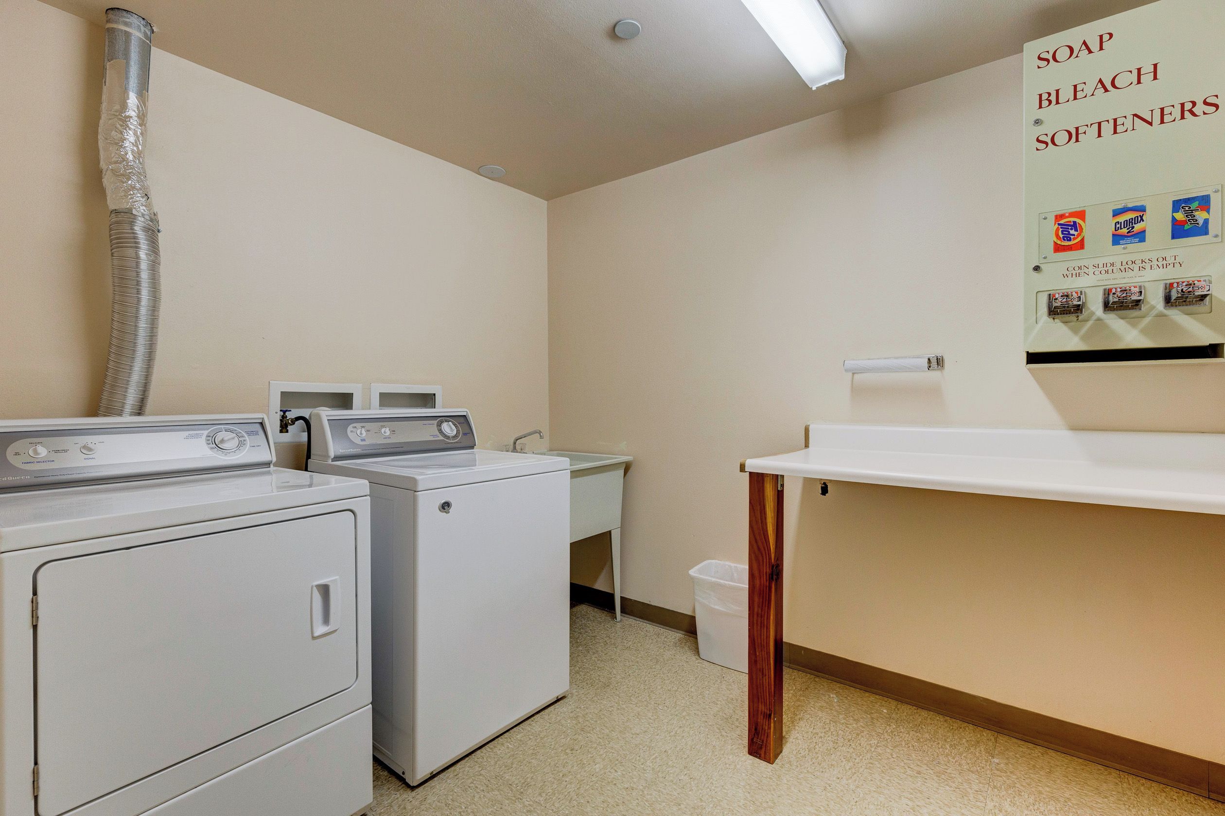 A laundry room with two washing machines, a sink, and a bulletin board with soap and bleach softener advertisements.