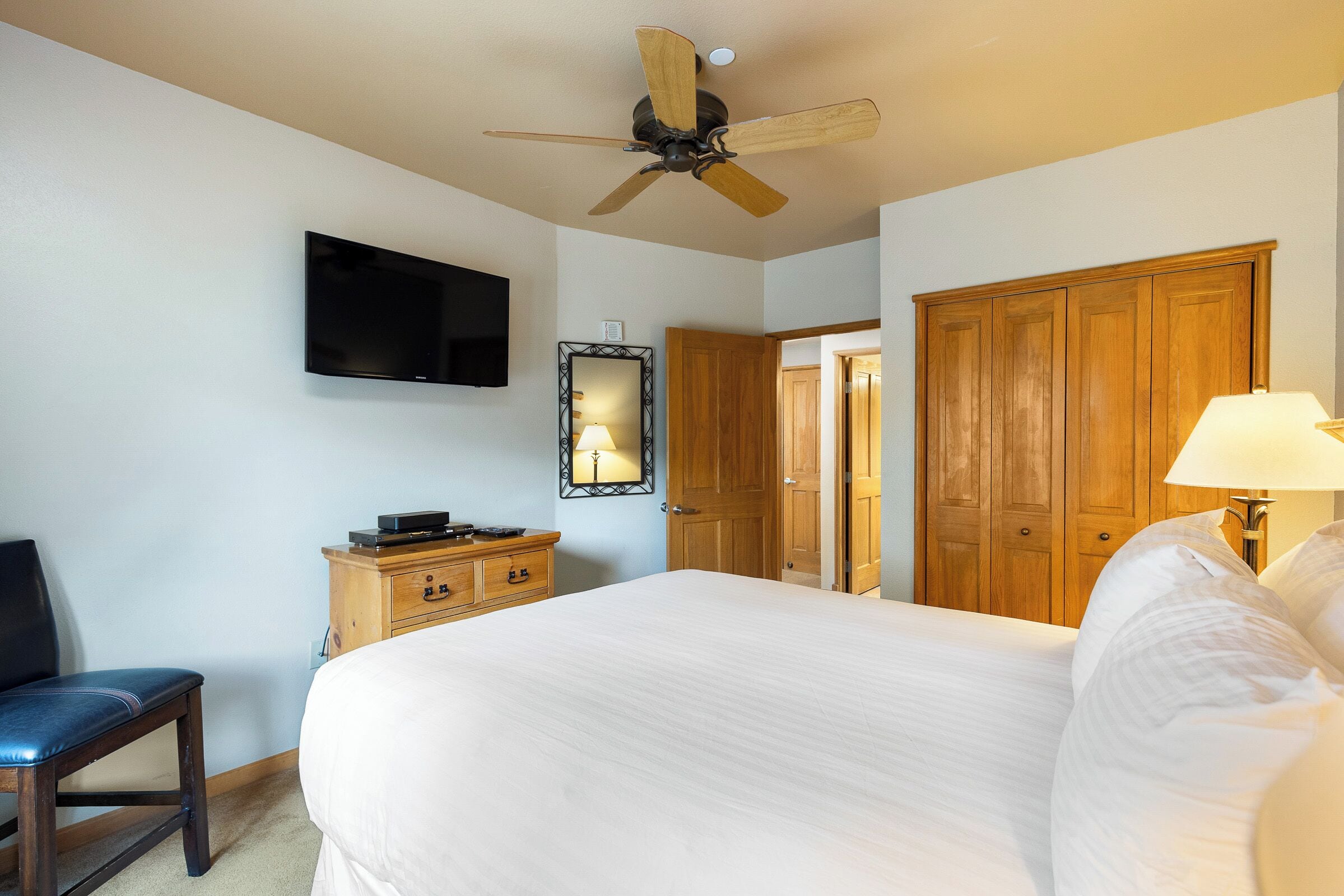A hotel room with a bed, a television mounted on the wall, a ceiling fan, wooden furniture, and a mirror.