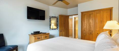 A hotel room with a bed, a television mounted on the wall, a ceiling fan, wooden furniture, and a mirror.