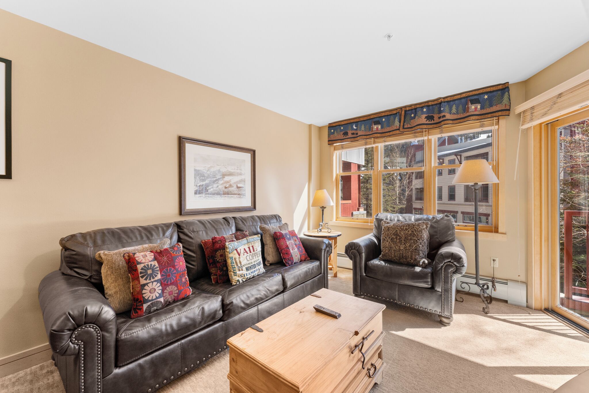 A living room with a brown leather sofa, a wooden coffee table, and a black leather armchair. There is a framed picture on the wall and a window with a view of buildings.