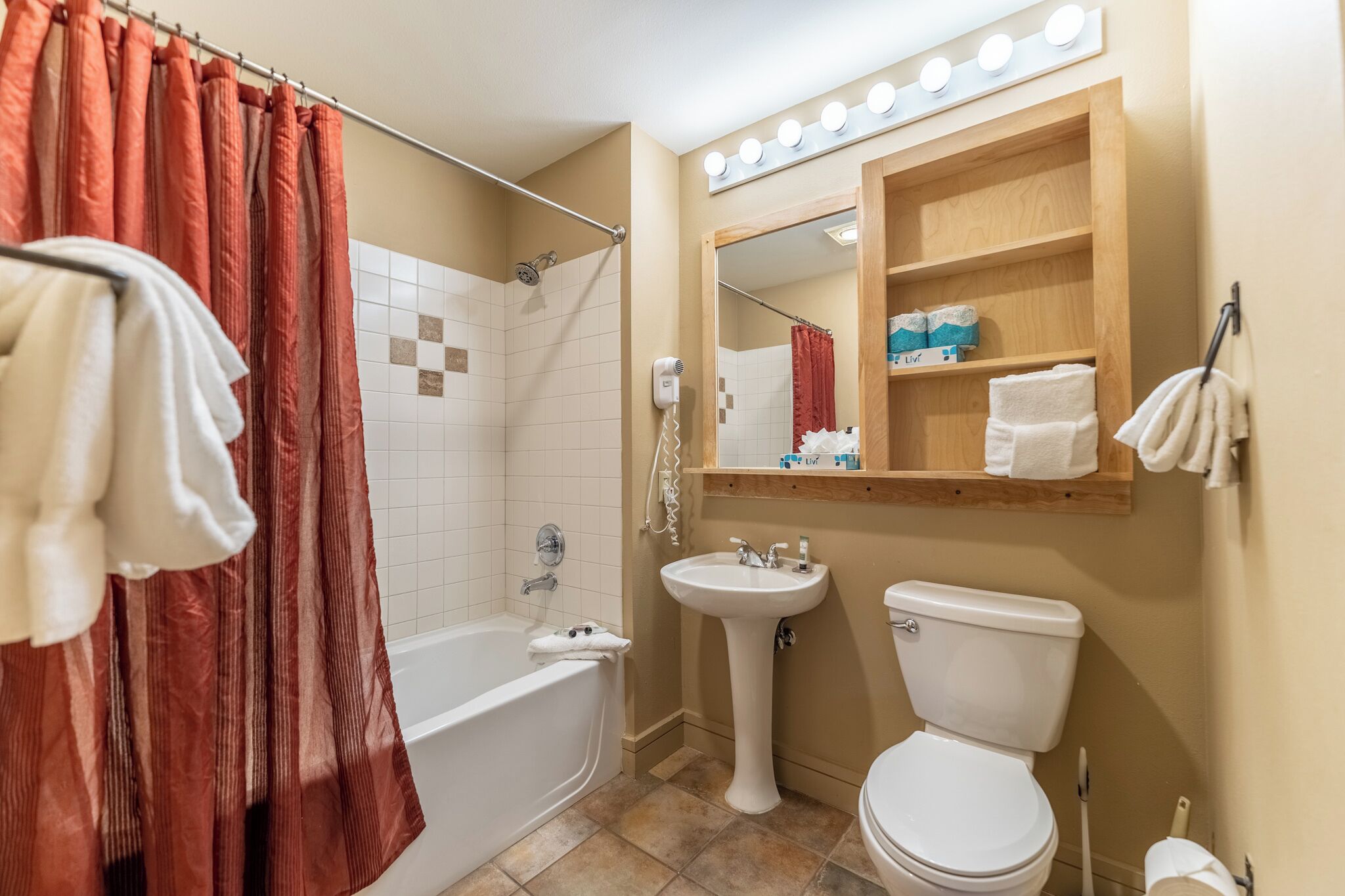 Un baño con bañera, lavabo e inodoro. Hay una cortina de ducha, un tocador con espejo y un estante con artículos de aseo.