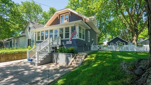 A two-story house with a front porch, a flag, and a white picket fence.