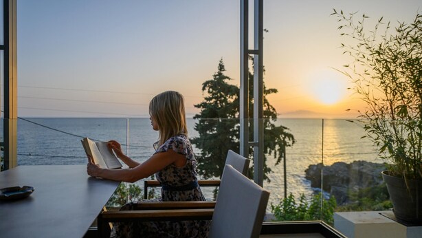 A person reading a book at a table with a view of the sea and trees at sunset. - Petra Deluxe Studio Suite (West Mani)