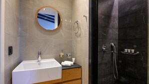 A bathroom with a white sink, wooden cabinet, and a round mirror. There is a shower with a dark tiled wall and a handheld showerhead. - Petra Deluxe Studio Suite (West Mani)