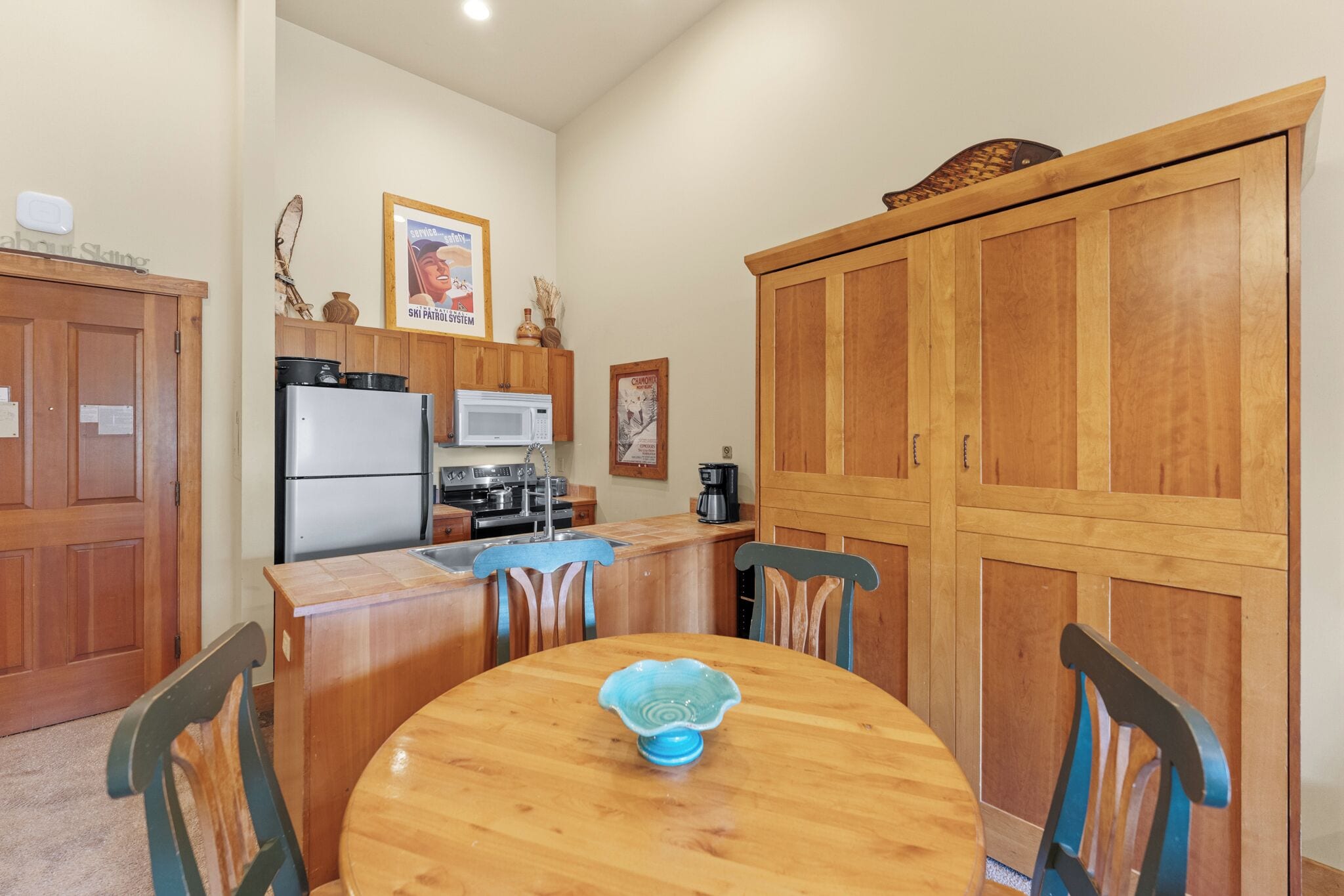 A compact kitchen with wooden cabinets, a refrigerator, microwave, and a dining table with chairs.