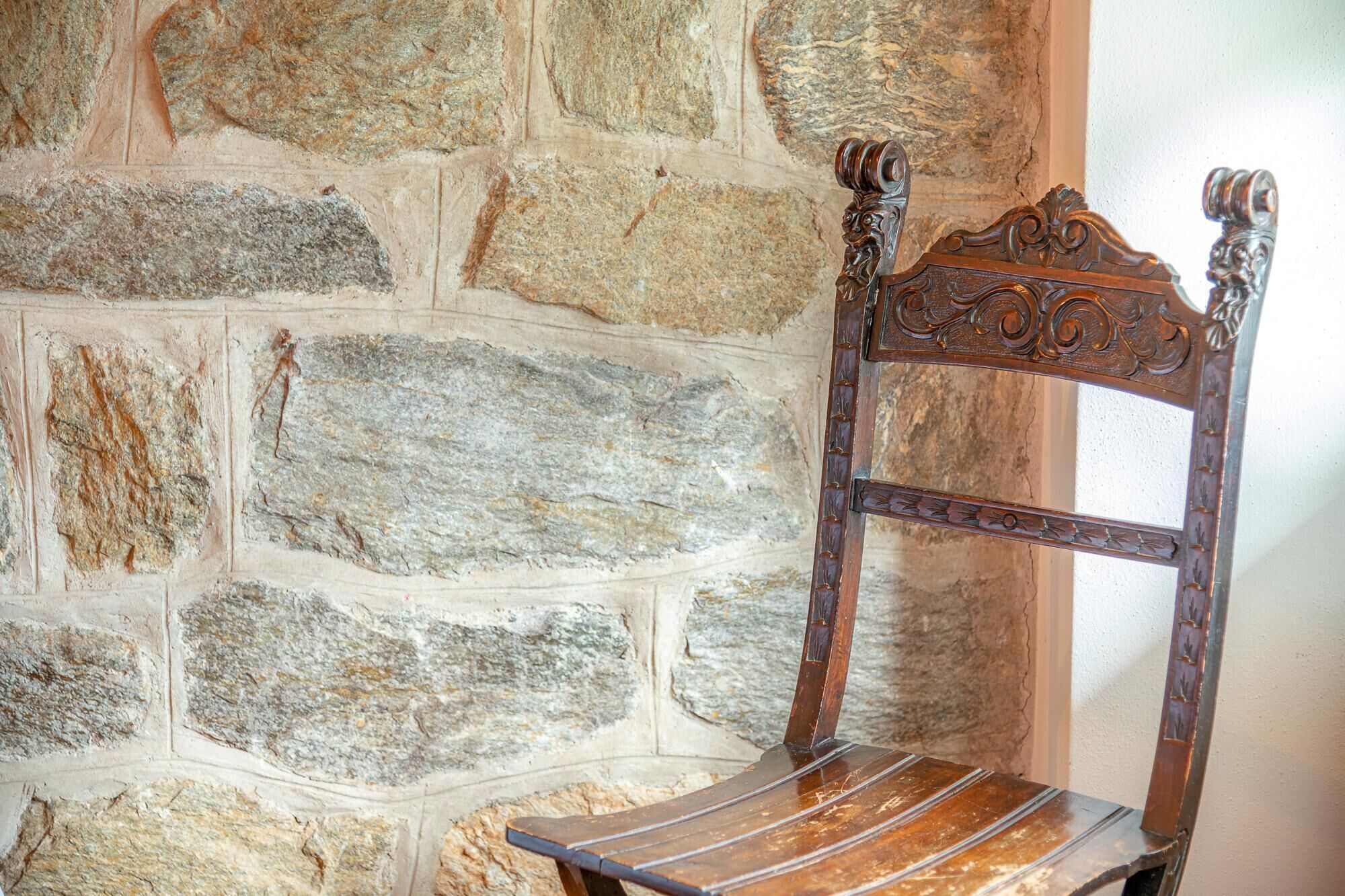 A wooden chair with a carved backrest and a slatted seat, positioned against a stone wall.