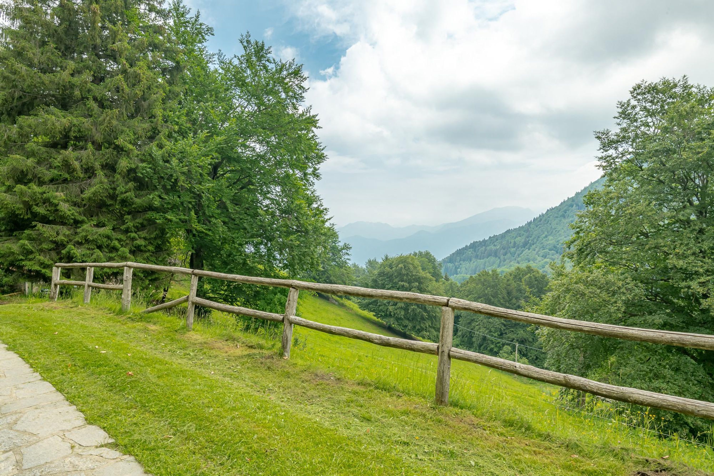 Ein gepflasterter Weg führt zu einem Holzzaun mit Blick auf ein üppiges, grünes Tal und entfernte Berge.