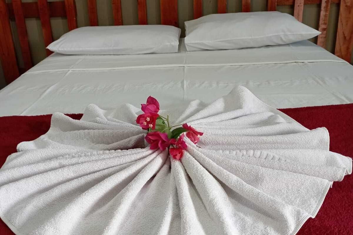 Una cama bien tendida con sábanas blancas y una manta roja, un cabecero de madera y una sola flor rosa sobre el edredón.