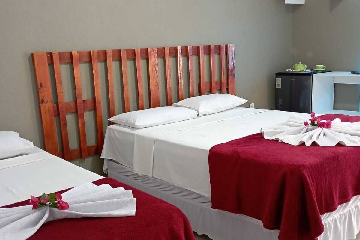 Habitación de hotel con dos camas, cada una con ropa blanca y mantas rojas, cabecero de madera, una kitchenette pequeña y un equipo de aire acondicionado instalado en la pared.