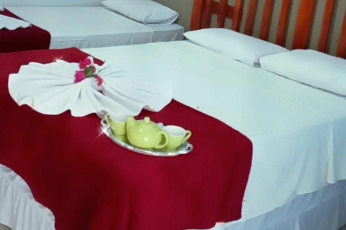 Habitación de hotel con dos camas, una colcha roja y blanca, un almohadón blanco y una tetera verde sobre un mantel rojo.
