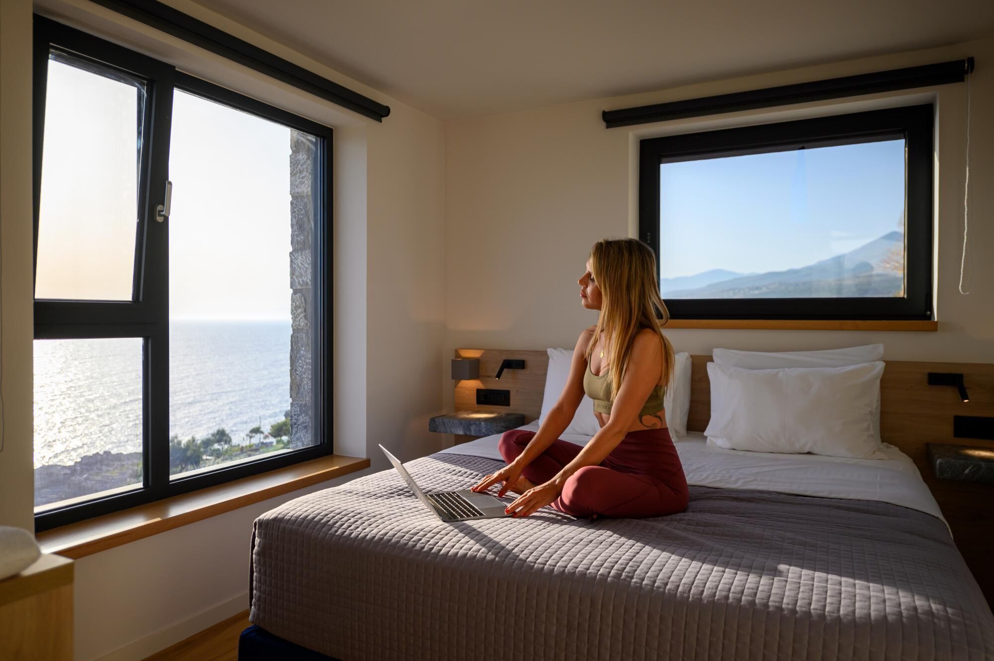 Una mujer sentada en una cama en una habitación con ventanas amplias que dan a un cuerpo de agua y montañas.