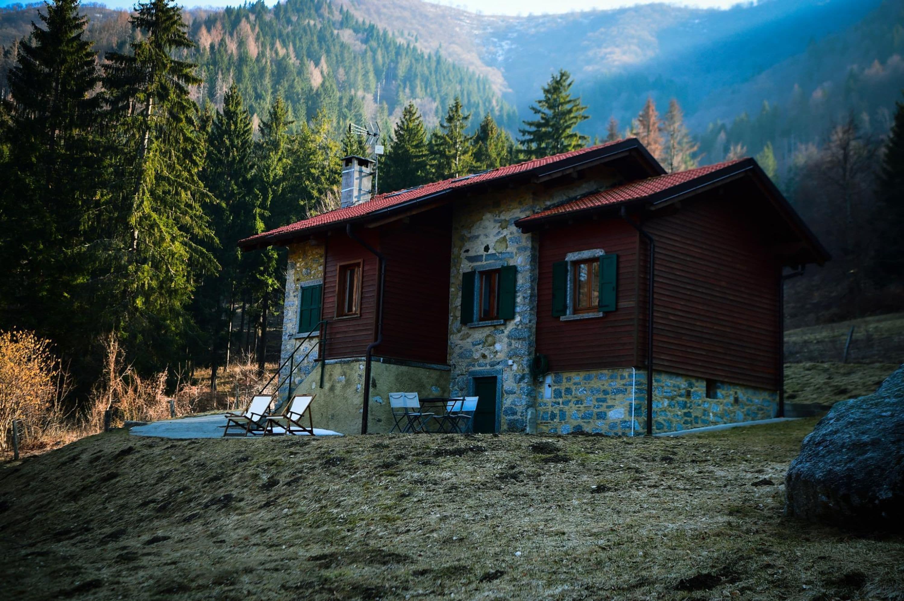 A rustic cabin with a stone facade and wooden exterior, situated in a forested area with a view of mountains in the background.