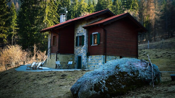 A rustic cabin with a stone foundation and wooden exterior, situated in a forested area with a large rock in the foreground. - Il Faggio by Wonderful Italy (Bellagio)