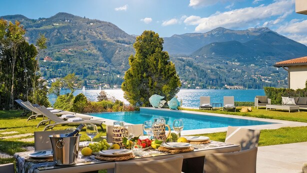 A poolside dining area with a view of a lake and mountains, lounge chairs, and a table set for a meal. - Villa Bianca (San Felice del Benaco)
