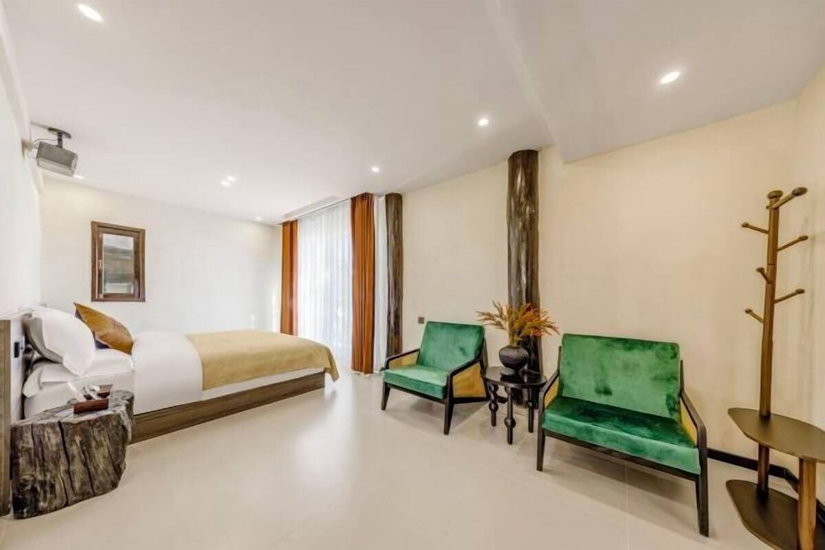 Una habitación de hotel con una cama, dos sillones verdes, una mesita y un perchero.