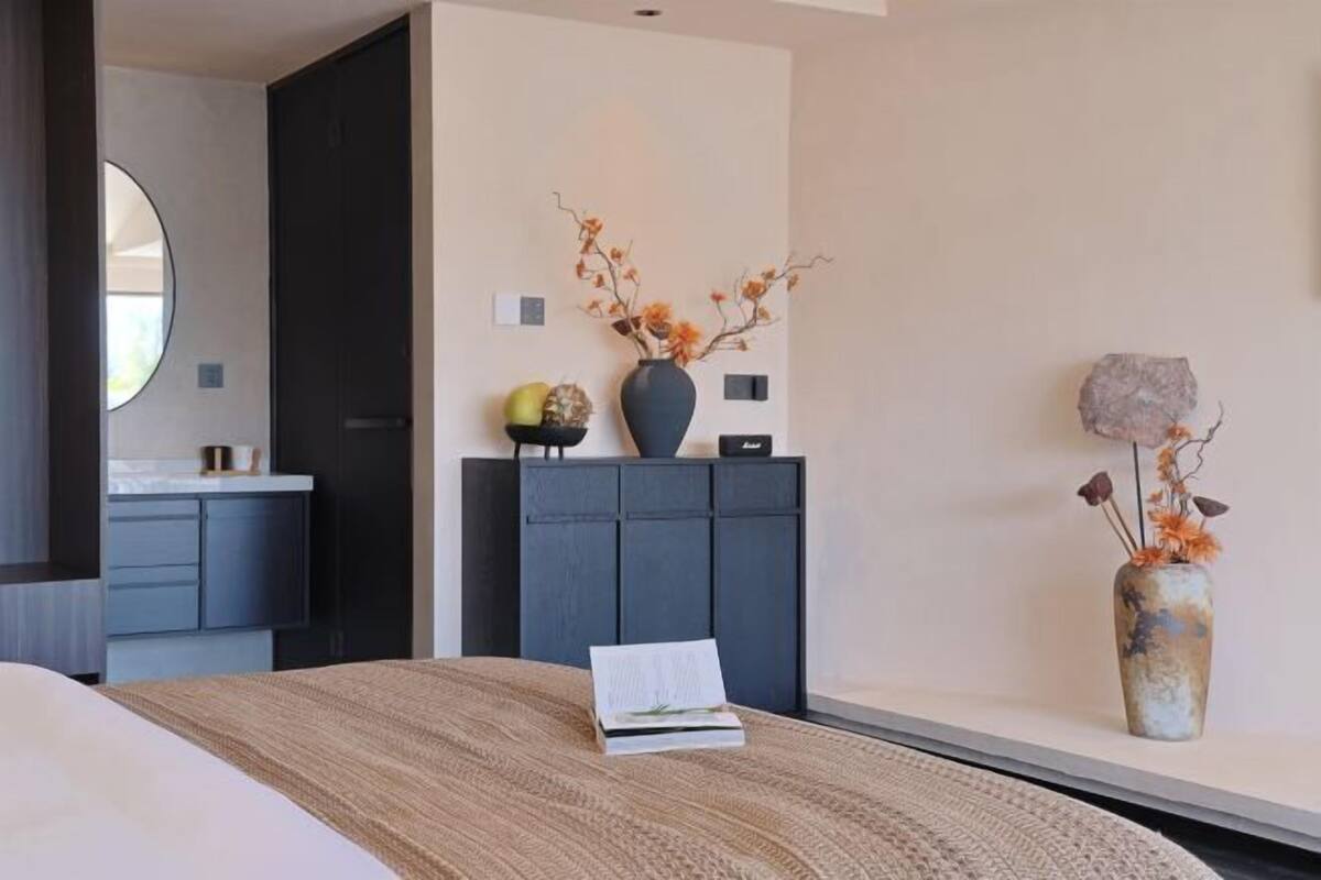 Una habitación de hotel moderna con una cama, un aparador de color oscuro, un florero con flores secas y un espejo redondo.