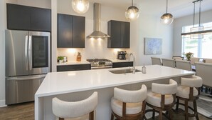 Cozinha moderna com ilha central, eletrodomésticos de aço inoxidável, iluminação pendente e área de refeições com banquetas.