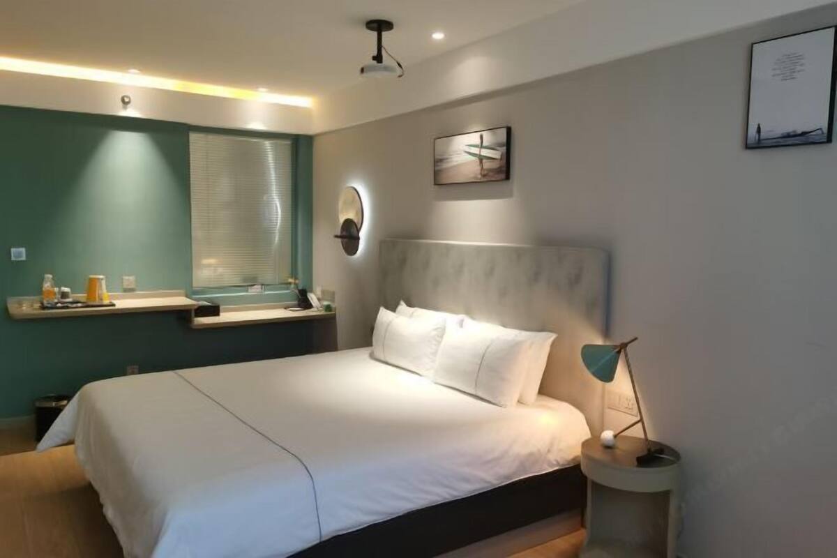 Ein modernes Hotelzimmer mit einem großen Bett, einem Nachttisch mit Lampe, einem Fenster mit Jalousien und einem an der Wand hängenden Kunstwerk.