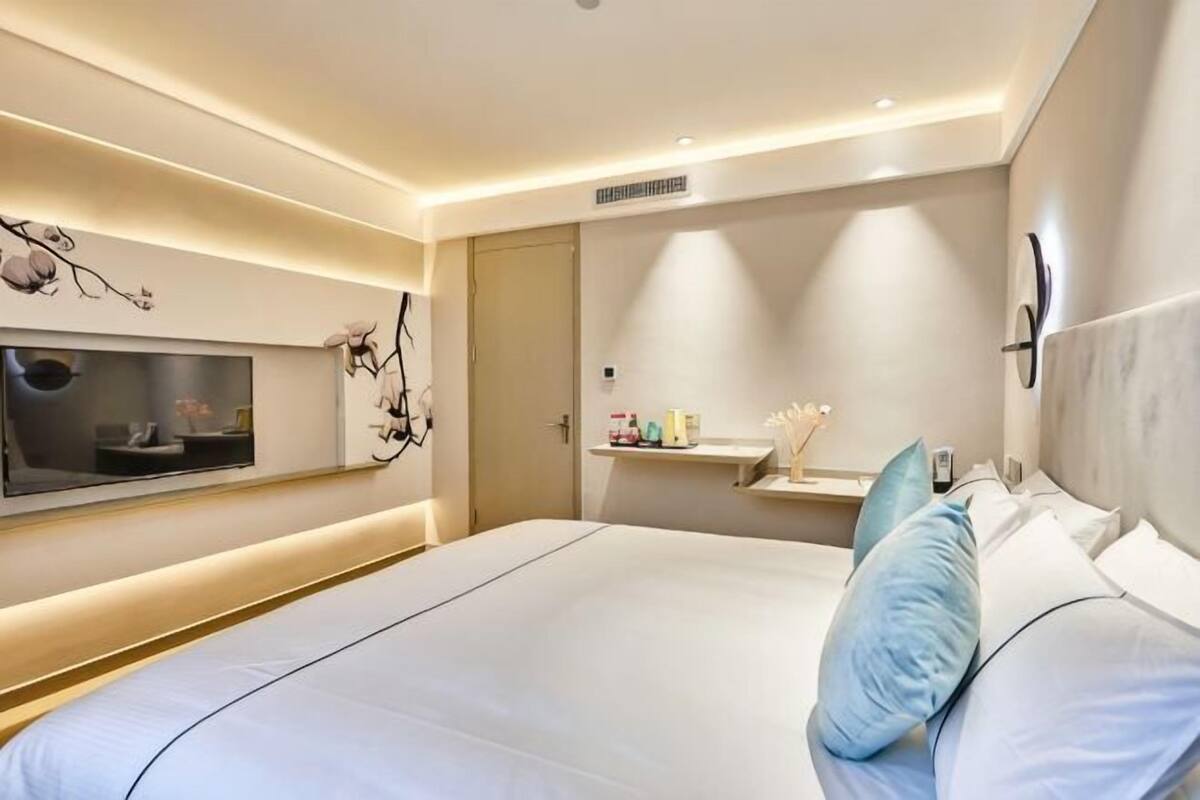 Ein modernes Hotelzimmer mit einem großen Bett, einem Fernseher, einem Schminktisch mit Spiegel und einem an der Wand befestigten Regal.