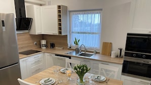 A modern kitchen with white cabinetry, a stainless steel refrigerator, a built-in oven, and a dining area set for four with white chairs and a wooden table. - Diamentowa 7 Rent Home (gmina Dopiewo)
