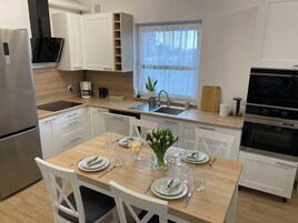 Une cuisine moderne avec des meubles blancs, un réfrigérateur en acier inoxydable, un four encastré et un coin repas aménagé pour quatre personnes, avec des chaises blanches et une table en bois.
