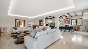 A spacious living area with a large sofa, coffee table, and dining table. There are multiple seating options and a kitchen area visible in the background. - Villa Xula - Marbella Club (Marbella)