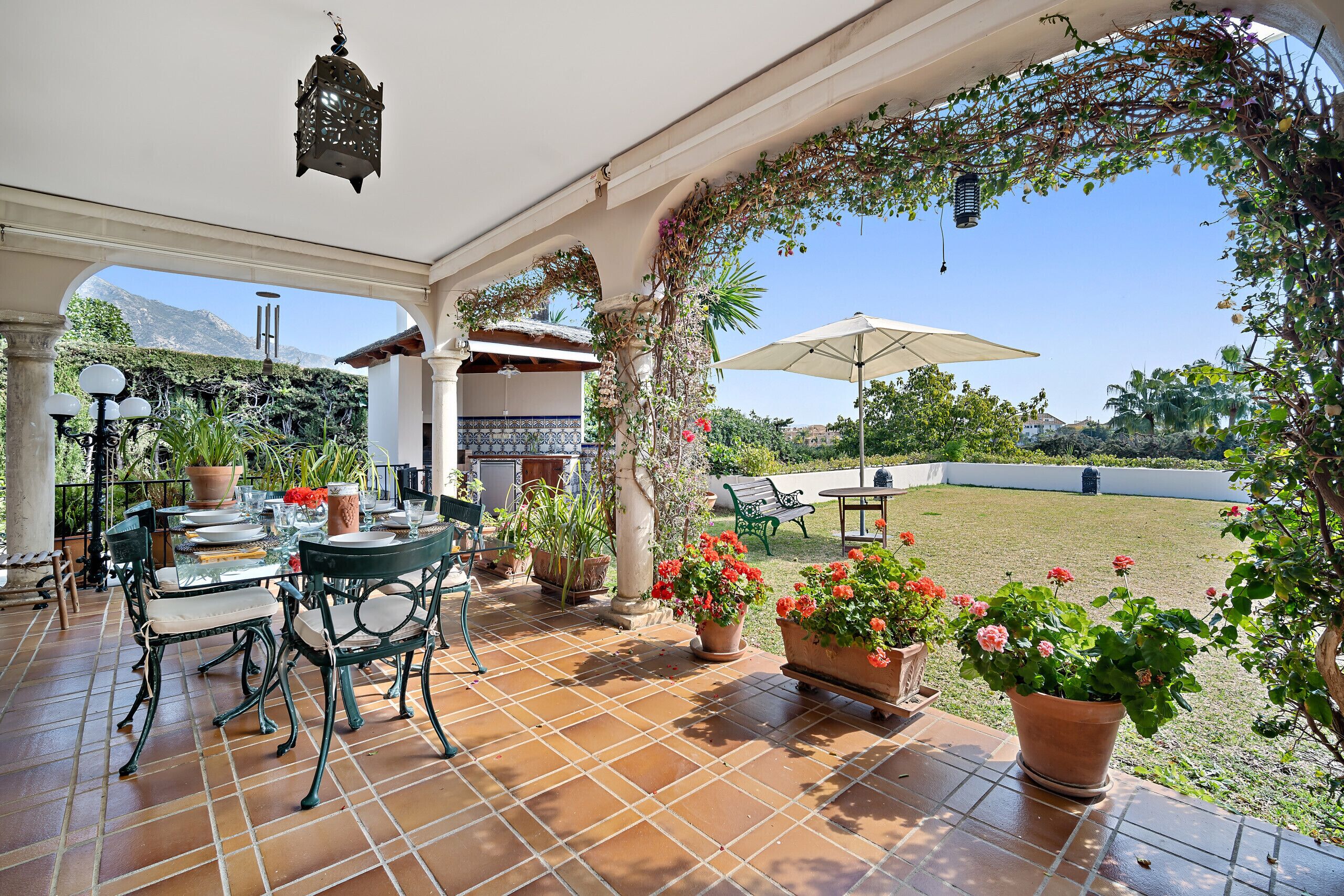 An outdoor patio with a tiled floor, a table with chairs, potted plants, and a view of a grassy area and mountains in the background.