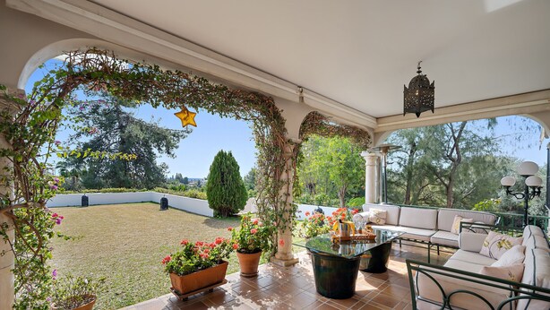 A covered patio area with a glass table, outdoor seating, potted plants, and a view of the garden. - Villa Xula - Marbella Club (Marbella)