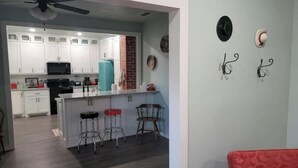 Une cuisine moderne avec des armoires blanches, un micro-ondes noir et un distributeur d’eau turquoise. On y trouve trois tabourets près de l’îlot central et un banc rouge avec coussin, placé au premier plan.