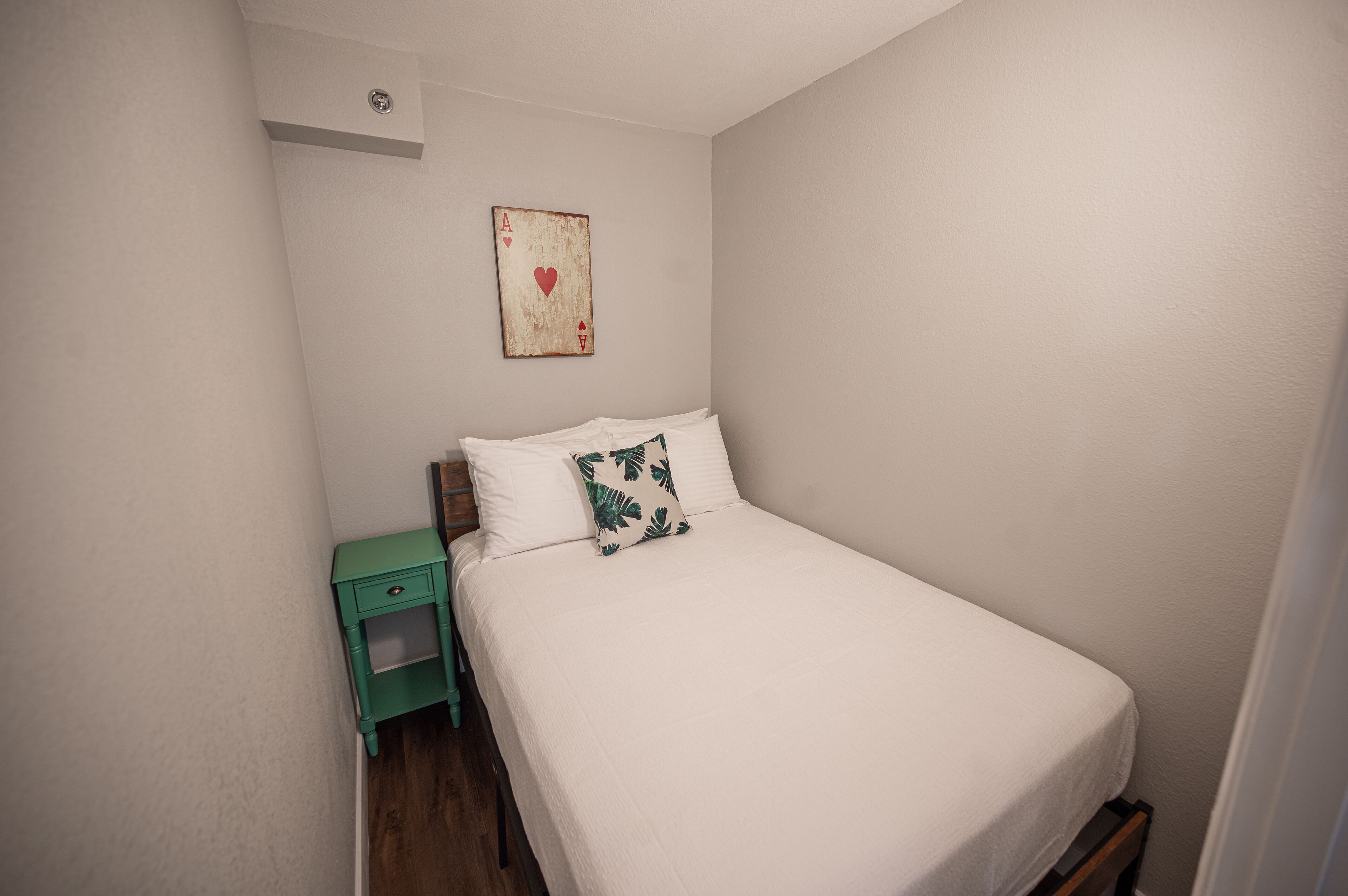 A small, neatly made bed with white bedding and a single decorative pillow. A small green nightstand with a drawer is positioned to the left of the bed.