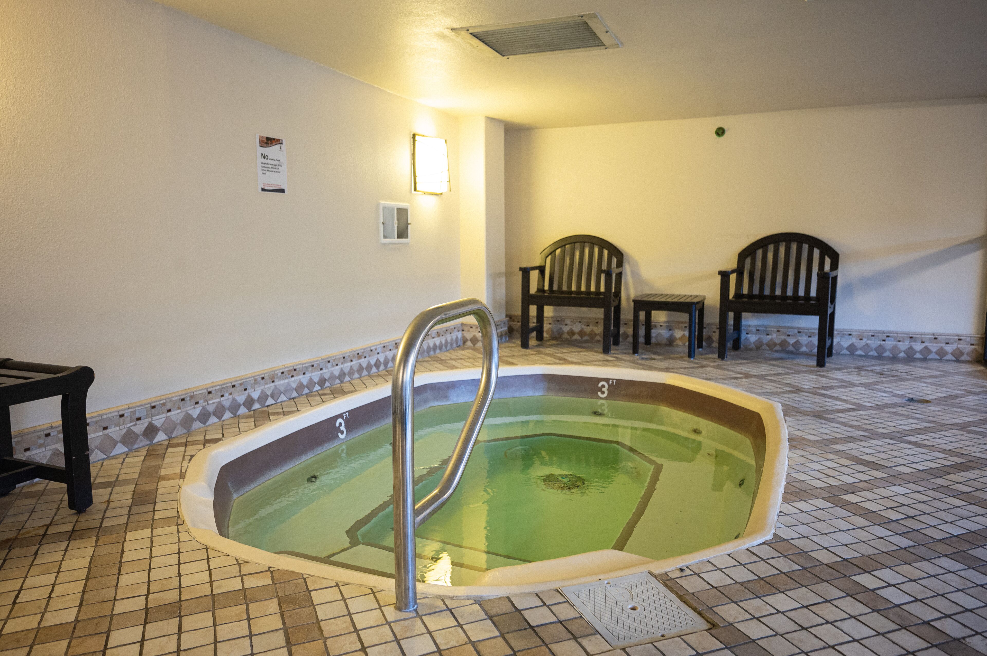 A small indoor pool with a metal ladder, two wooden chairs, and a tiled floor.