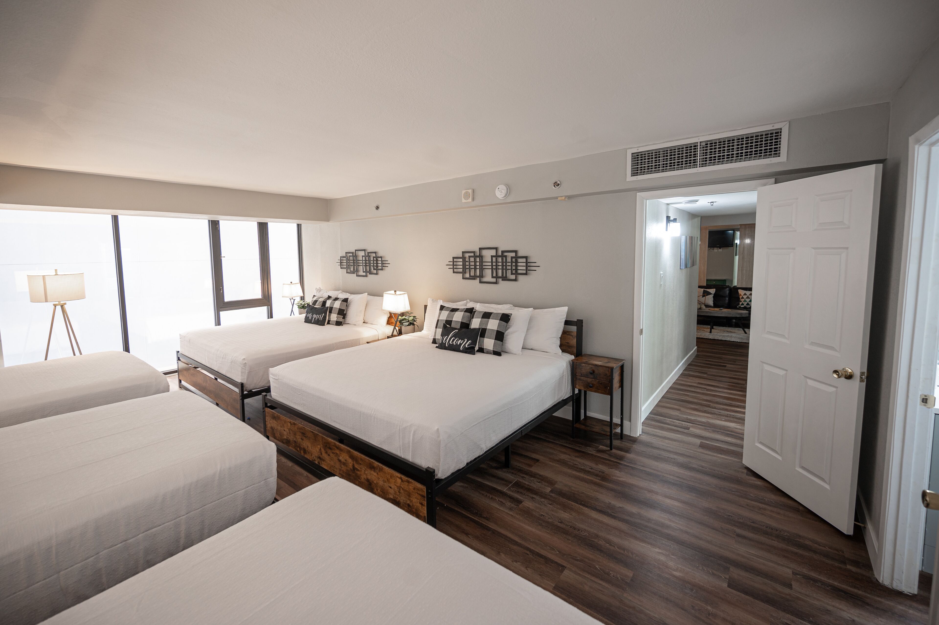 A hotel room with two beds, a bedside table with a lamp, a wall-mounted air vent, and a door leading to another room.