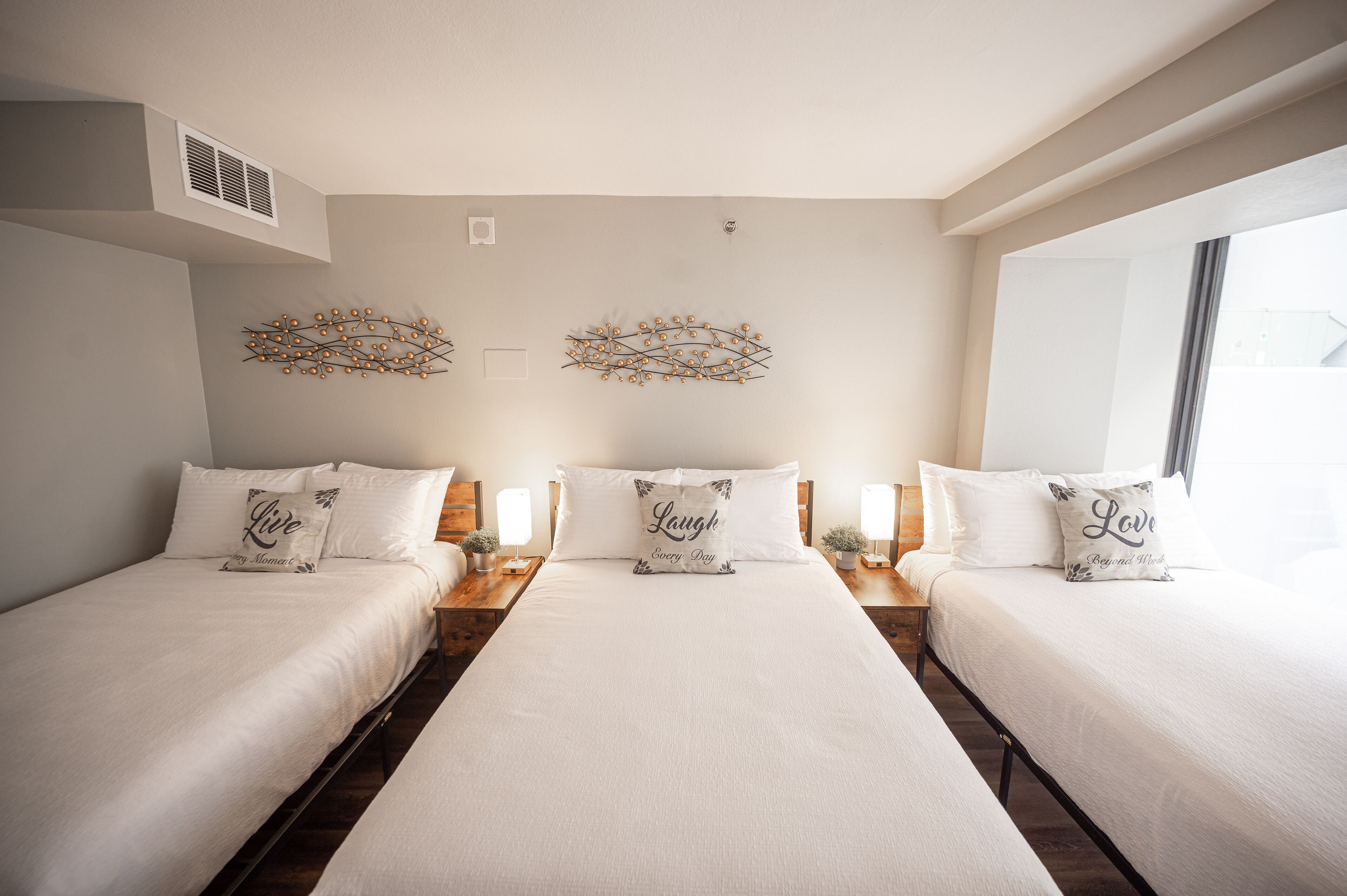 A hotel room with three beds, each with a white bedspread and matching pillows. There are wall-mounted light fixtures and a vent on the wall.