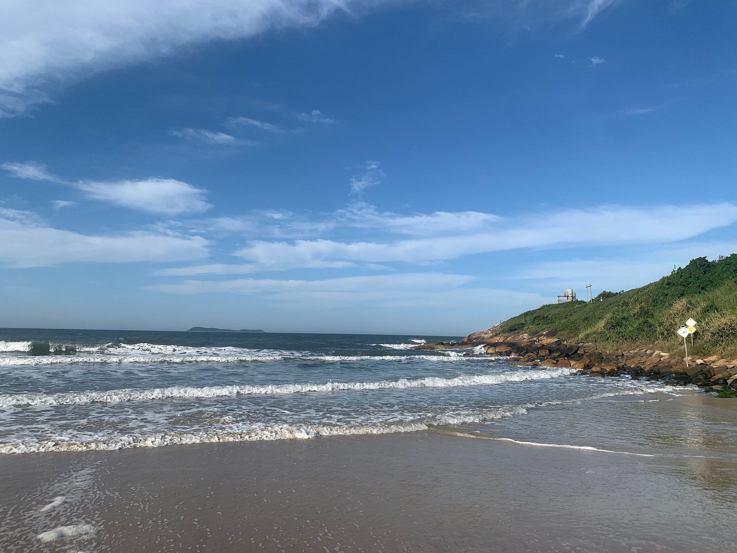 Una playa con olas, un litoral rocoso y una colina con vegetación.