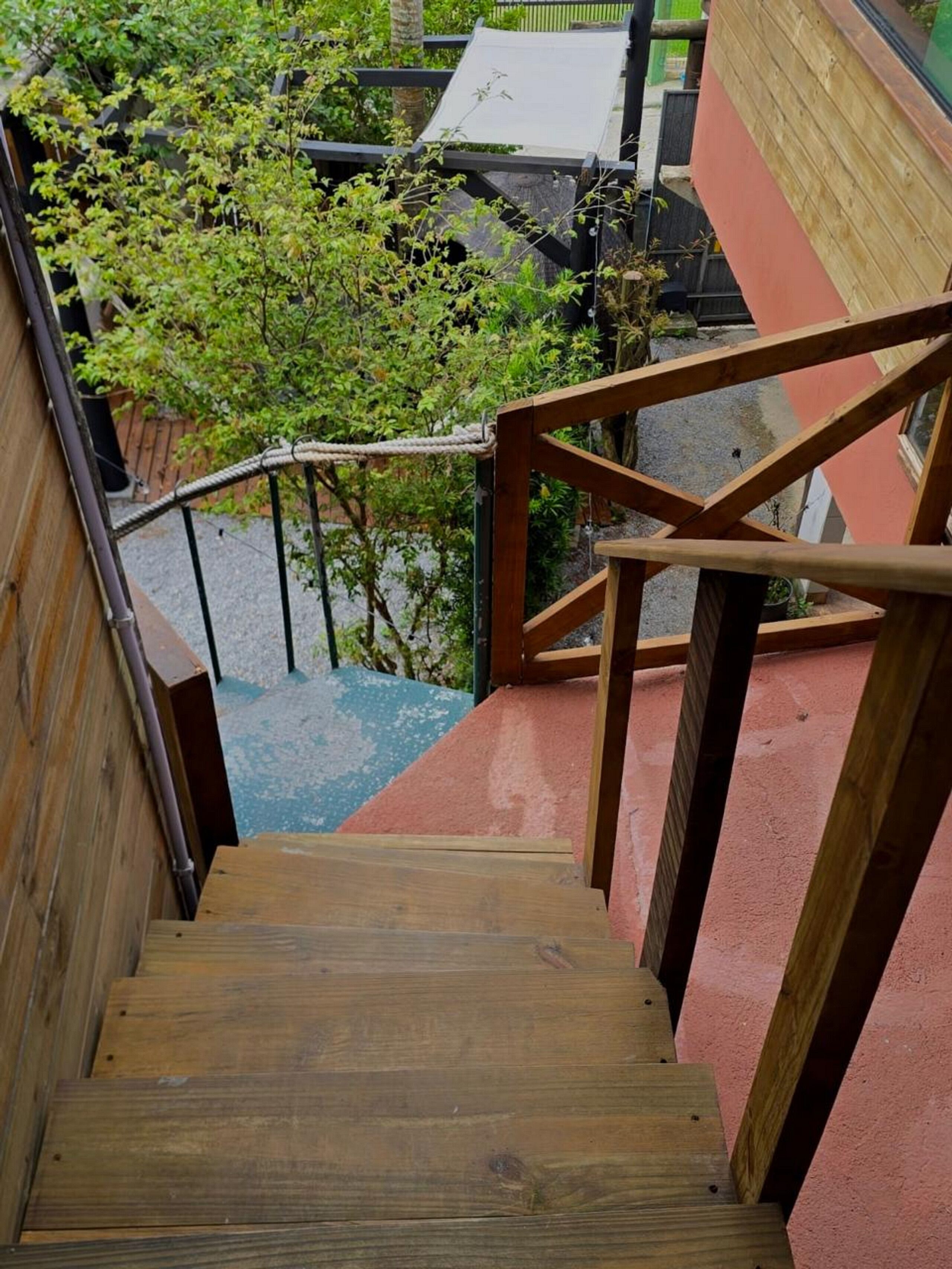 A wooden staircase with a metal handrail leading to a red-tiled area with a view of greenery and buildings.