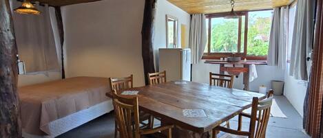 A rustic wooden room with a large dining table, chairs, a bed, and a window with curtains.