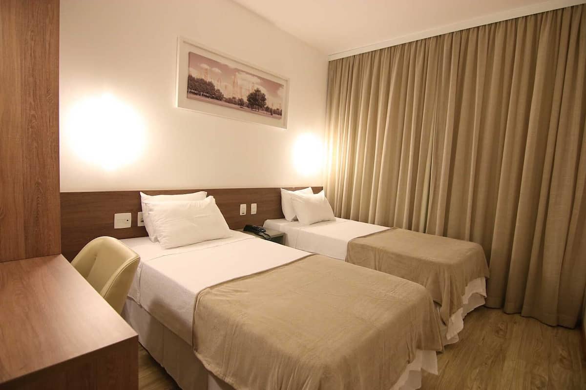 Habitación de hotel con dos camas, un escritorio, una silla y un ventanal grande con cortinas.