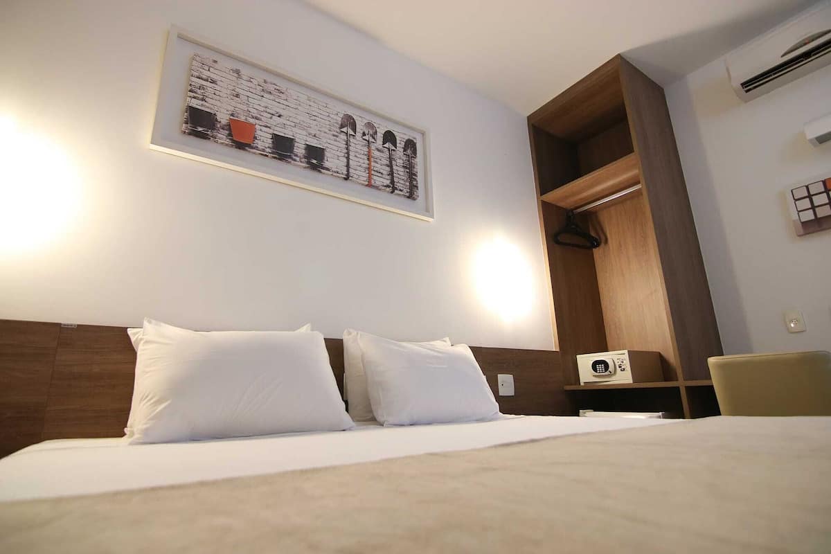 Habitación de hotel con cama, perchero, un cuadro enmarco y una mesita con un radio.