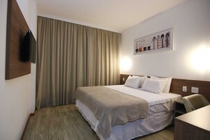 Quarto de hotel com cama, televisão de tela plana na parede, quadro sobre a cama e escrivaninha com cadeira.