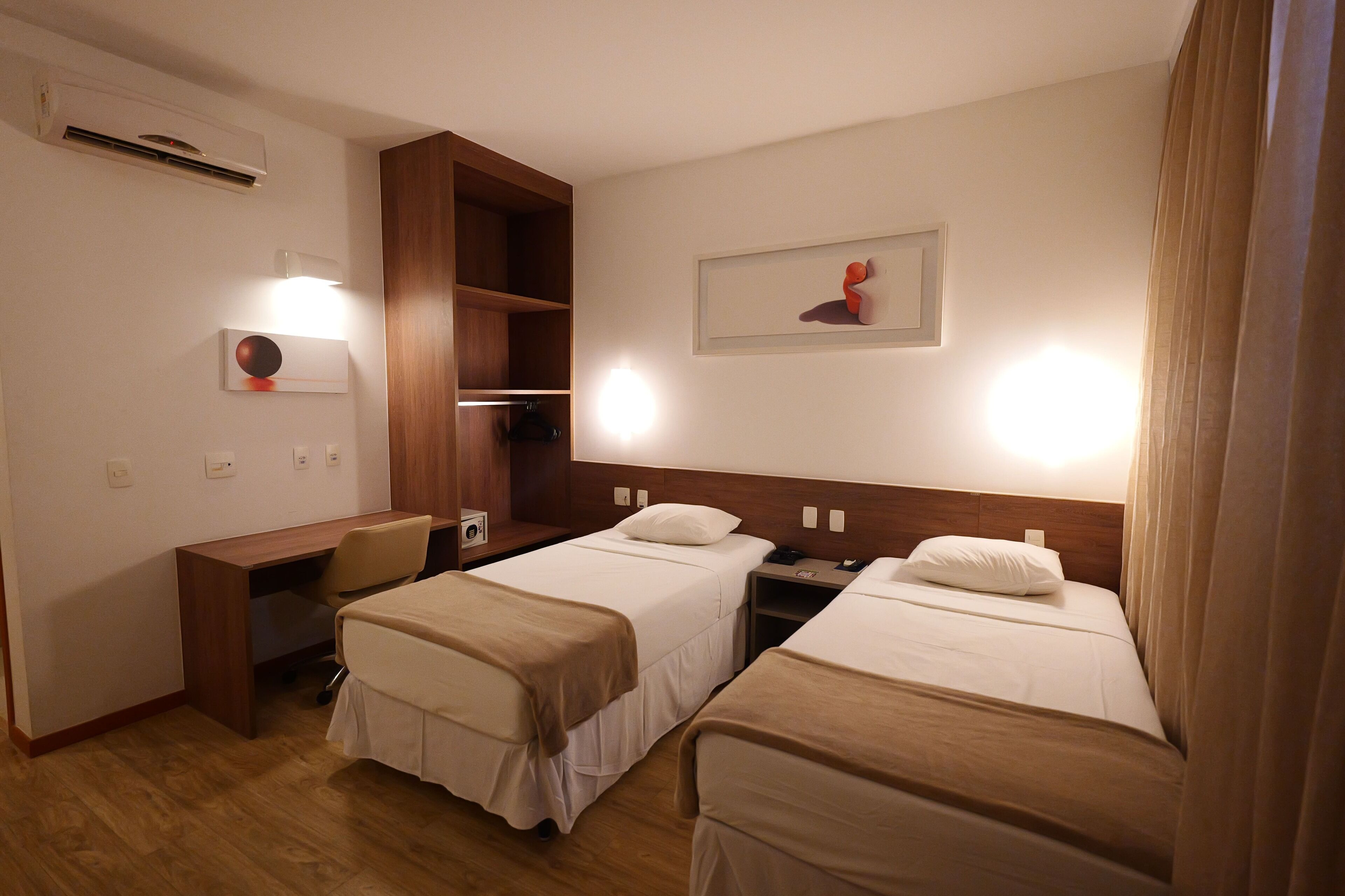 A hotel room with two single beds, a desk, a chair, and a wooden wardrobe.