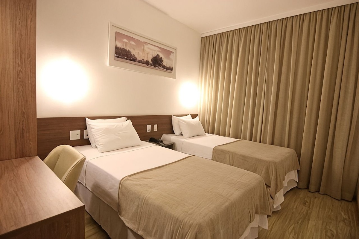A hotel room with two beds, a desk, a chair, and a large window with curtains.
