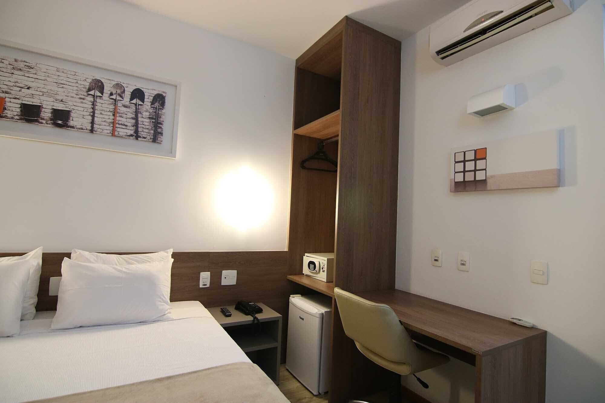 A hotel room with a bed, a desk, a chair, a small fridge, a phone, and a wall-mounted air conditioner.