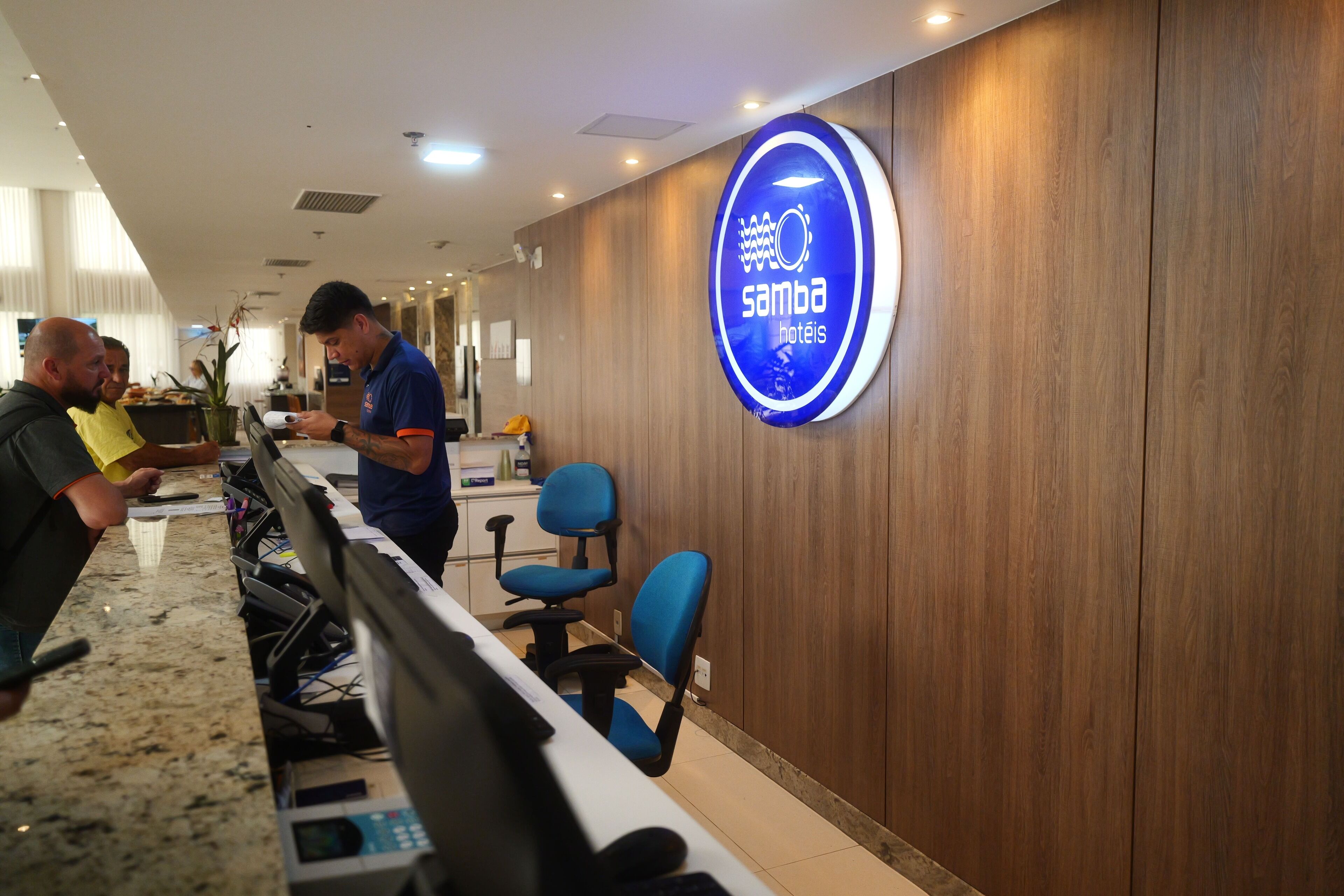 Una zona de recepción con pared de madera, un letrero azul que dice "Samba Hotels" y un mostrador con computadoras y sillas.