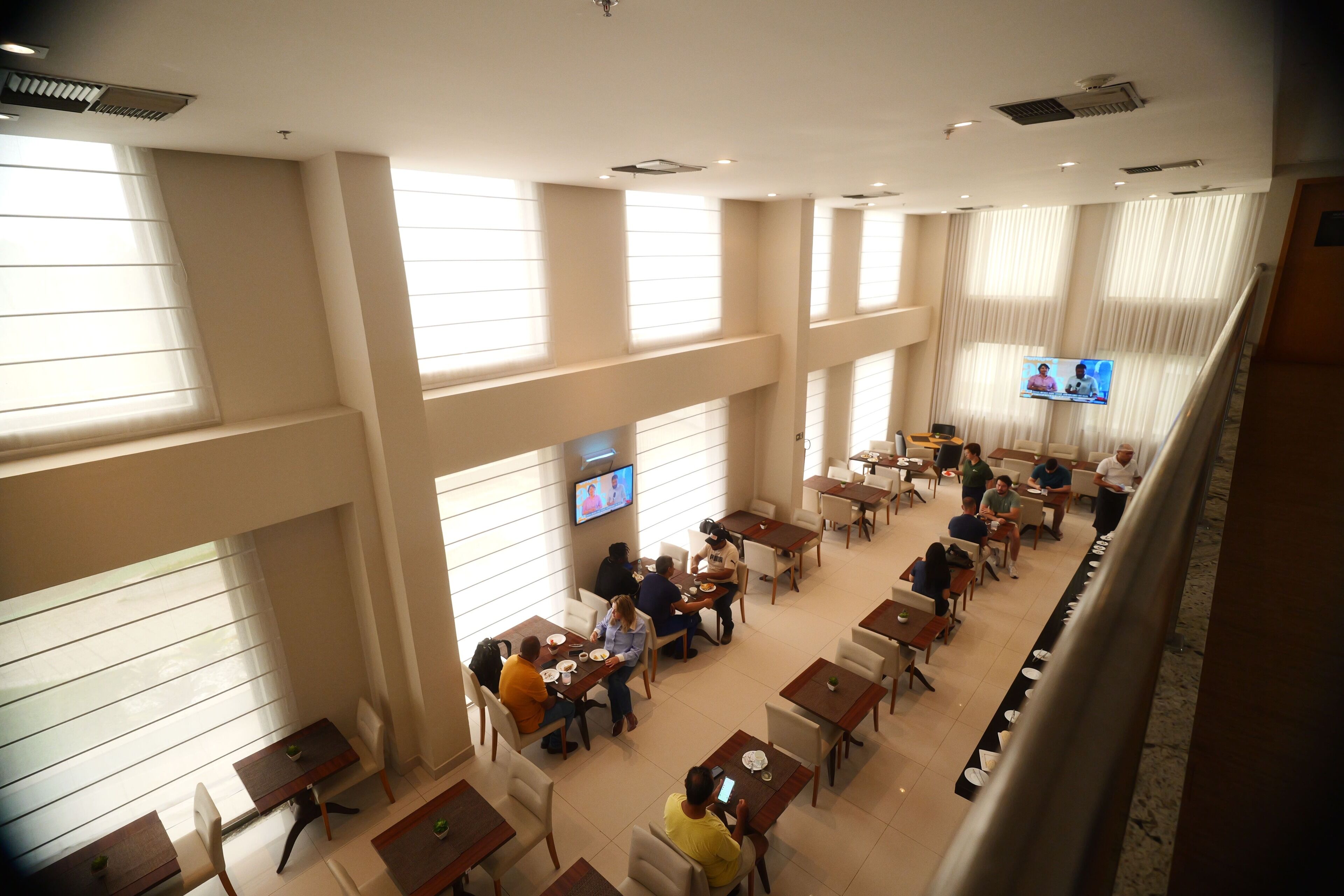 Un vaste espace dédié à la salle à manger, avec de nombreuses tables et chaises, de grandes fenêtres équipées de stores et un écran de télévision diffusant une émission.
