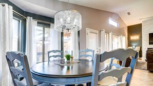 A dining area with a round table, six chairs, a chandelier, and large windows with curtains.