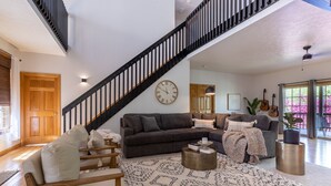 Un vaste salon comprenant un canapé marron foncé, un canapé marron clair et un tapis à motifs. Un escalier avec des rampes noires mène à l’étage supérieur.