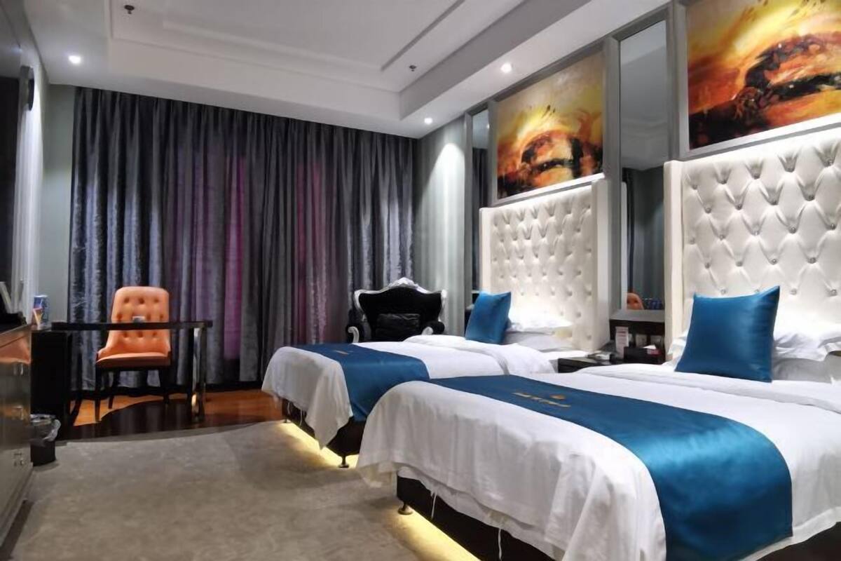 A hotel room with two beds, a desk, an orange chair, and a large painting on the wall.