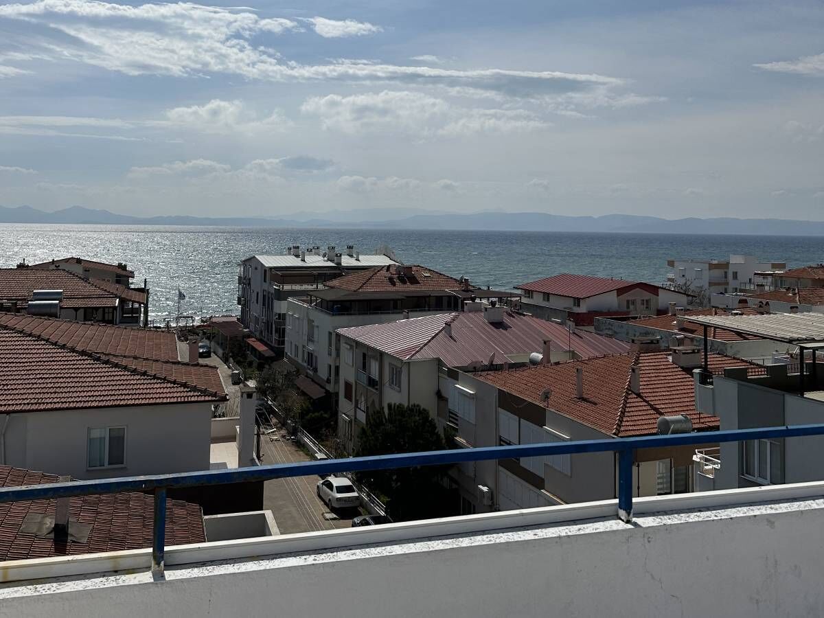 A coastal residential area with buildings featuring red-tiled roofs, a clear view of the sea, and distant mountains.