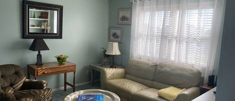 En stue med en ventilator, en beige sofa, en brun lænestol, et lille rundt bord med en bog og en lampe på et sofabord.