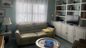A living room with a beige sofa, a coffee table with a book, a ceiling fan, and a bookshelf with various items. - Hatteras Village *** Southside 3 *** (Hatteras)